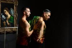 Two dancers wearing shimmery green, gold, and red clothing perform in front of a painting of Angel Gabriel by Carlo Dolci and a work by Joseph Liatela modeled after the railing in the PULSE nightclub. The background is a dark, matte black.